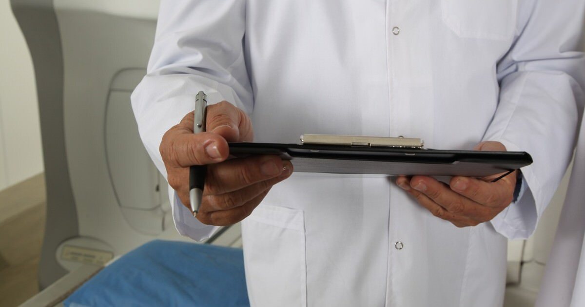 Medical professional in a white coat holding a clipboard and paperwork, with an empty patient bed visible in the background