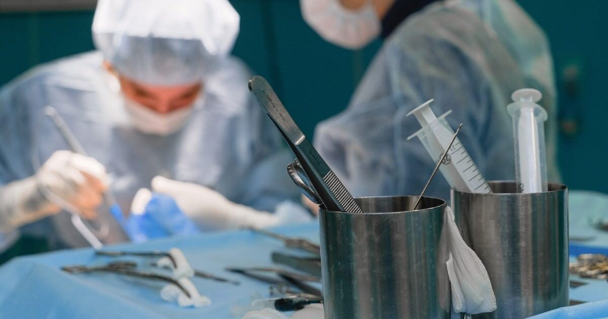 Tray of surgical supplies in the foreground, including syringes and metal tweezers, with surgeons working on a procedure in the background